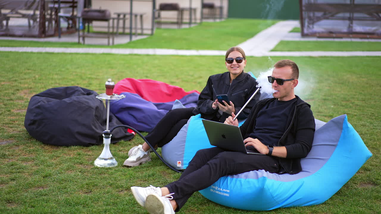Resting zone for good time outdoors. Couple in dark clothes and sunglasses sitting in bean bag chairs. People talk, use gadgets and smoke hookah.
