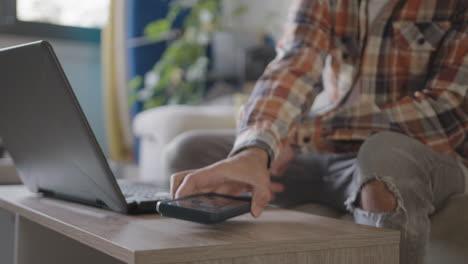 Una-Llamada-Entrante-Que-Distrae-A-Un-Joven-Blogger-Adulto-Que-Trabaja-Desde-Casa-En-Su-Sala-De-Estar-Usando-Una-Computadora-Portátil,-Distraído-De-La-Ventana-Emergente-De-Un-Teléfono-Móvil,-Tablas-De-Surf-En-El-Fondo