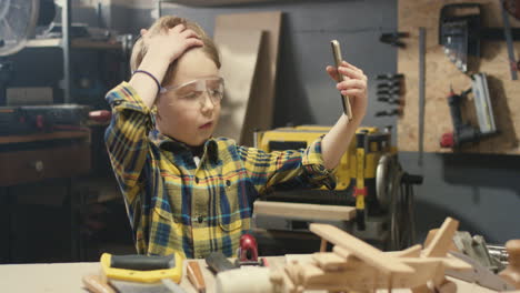 cute caucasian boy playing with wooden plane toy and making a selfie in carpentry workshop