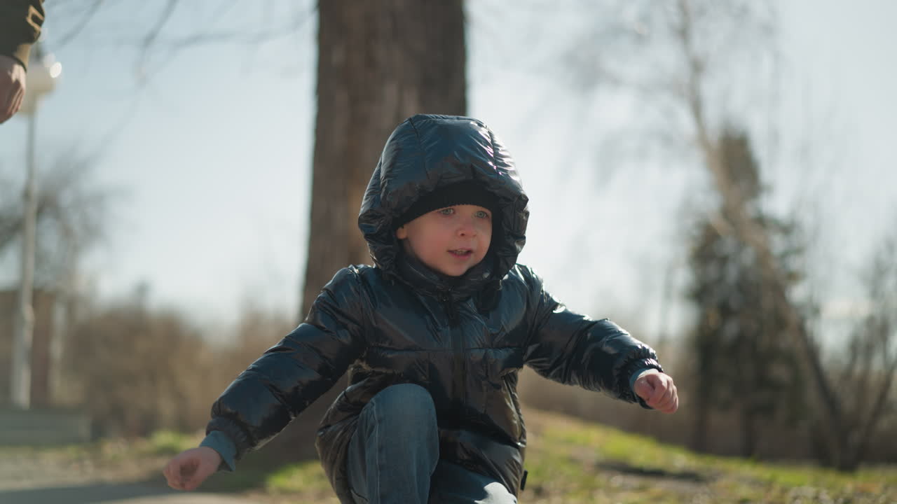 A close-up of a playful young boy squatting and moving around energetically, with his father, standing behind him, the boy is dressed warmly in a shiny black jacket and jeans
