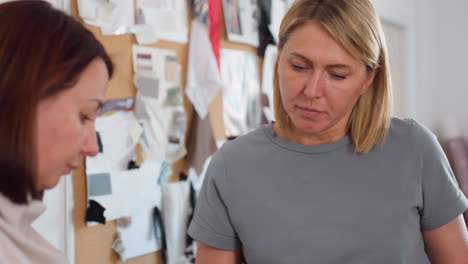 elegant women working together in creative studio focused on drawing project while one woman sketches on paper and other gives thumbs up approval, surrounded by inspiration boards and fabric samples