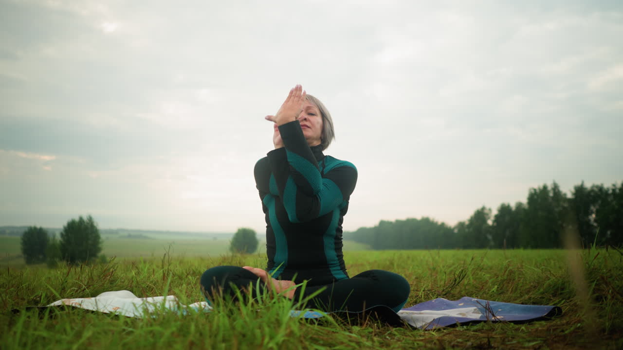 Middle-aged woman seated on yoga mat in misty grassy field, while practicing yoga with eyes closed cow face yoga, under a cloudy sky, surrounded by nature in a tranquil outdoor setting