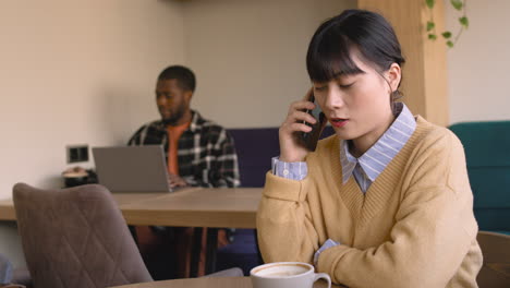 femme parlant au téléphone portable assise à table dans un café tandis qu'un homme travaillant sur un ordinateur portable derrière elle 1