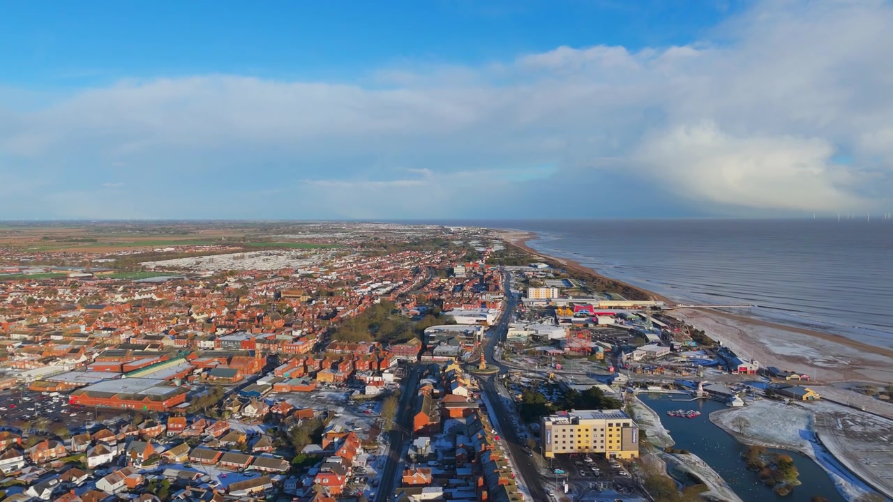 Flygfoto visar en kuststad med röda tak. Staden sträcker sig längs strandlinjen där havet möter sanden. Klar himmel med moln förstärker det pittoreska landskapet.