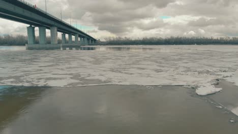 El-Agua-Del-Río-Medio-Congelada-Refleja-El-Puente-Gris-Y-Las-Nubes.