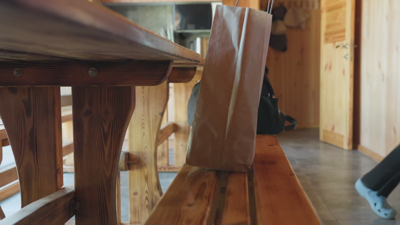 Hand in denim sleeve lifts shopping bag and black duffel from long wooden bench inside sunlit rustic kitchen cabin, low perspective framing warm pine planks, kettle and microwave