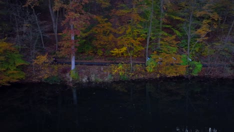 An-aerial-view-over-a-dark,-reflective-lake-in-the-morning-with-colorful-autumn-trees-on-the-bank