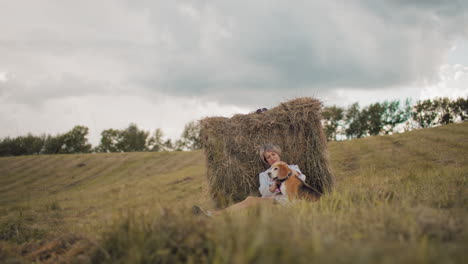 mujer de mediana edad con gafas de sol y traje casual se sienta contra la bala de heno en el campo, frotando suavemente la cabeza del perro, la hierba dorada, las colinas onduladas y el cielo nublado