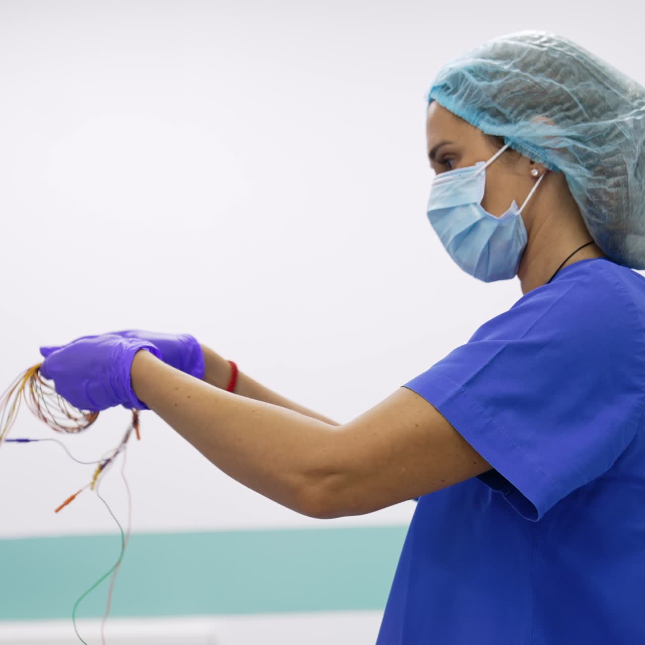 Profissional da saúde do sexo feminino usando uniforme azul, luvas, máscara e touca, desembaraçando os fios de um equipamento. Preparação para o procedimento de neuromonitoramento. Vista lateral