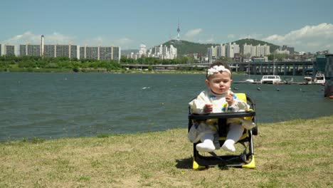 niña sentada mientras come al aire libre, hermoso río y edificios en el fondo - plano general