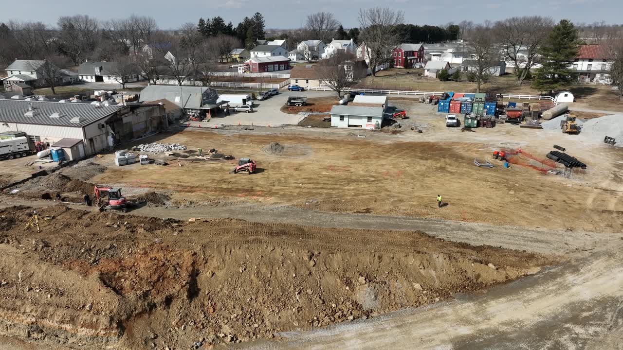 Heavy machinery operates at a construction site in a suburban environment. The landscape shows various building materials and vehicles, indicating ongoing site preparation.