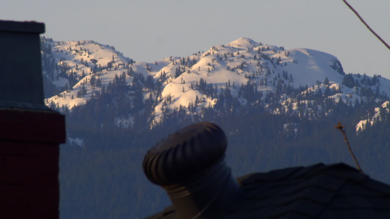 Spinning Roof Ventilation And Chimney Of A House In Vancouver, Canada With Snowy Mountain And Lush Pine Trees In The Background - Closeup Shot