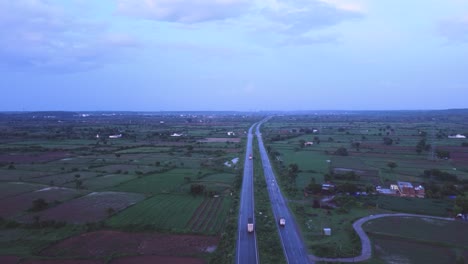 Toma-Aérea-De-Drones-De-Camiones-Transportadores-De-Mercancías-En-Una-Carretera-A-Través-De-Granjas-En-La-Zona-Rural-De-Gwalior-De-La-India-Durante-El-Crepúsculo.