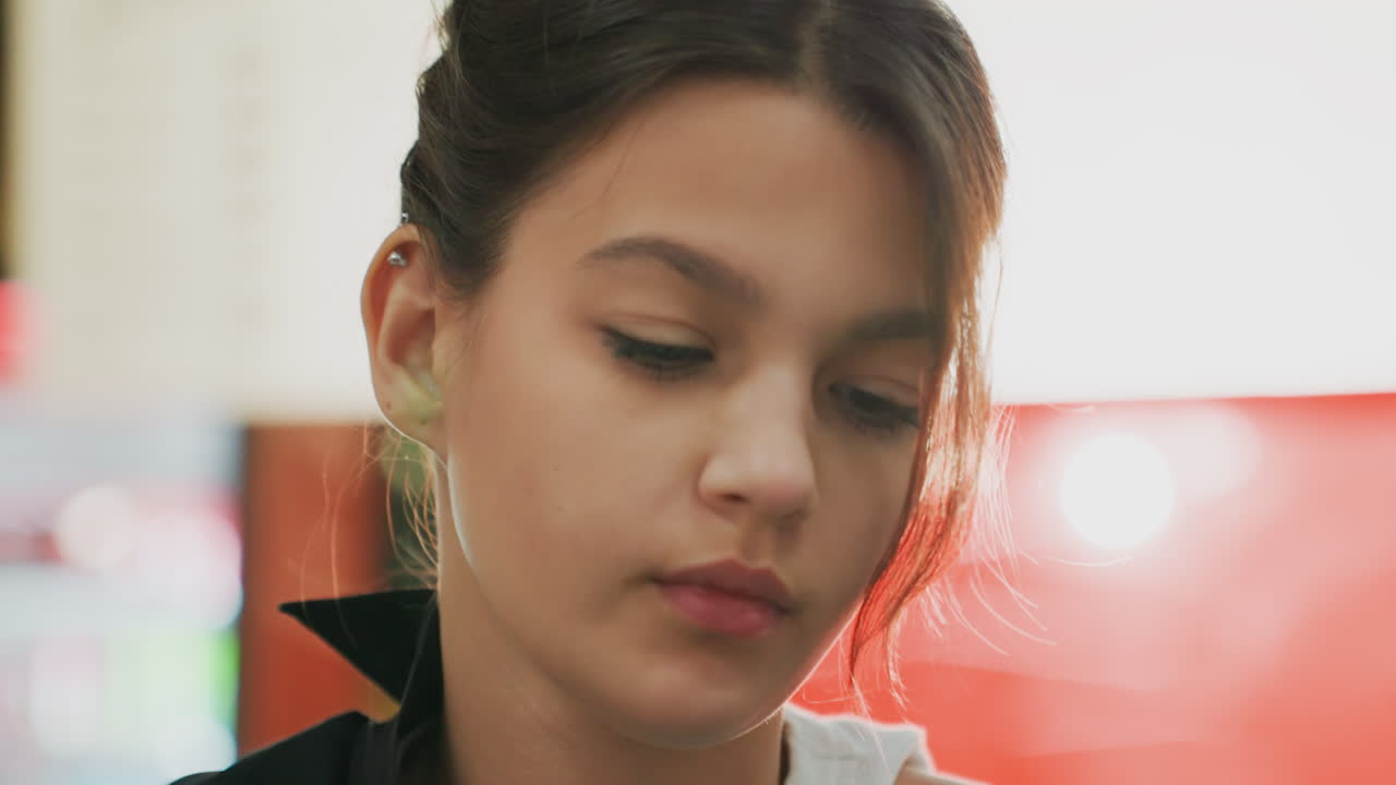 Una studentessa adolescente che si lega i capelli dietro le quinte, riflessa in uno specchio illuminato, con espressione concentrata mentre sistema lo chignon con una forcina e un braccialetto, soffusa retroilluminazione al neon, sequenza di un ritratto spontaneo che mostra la quieta preparazione alle prove, trucco leggero