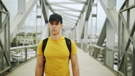 young attractive trendy young man wearing a cap and a backpack walking on a bridge on a sunny day with an urban city background