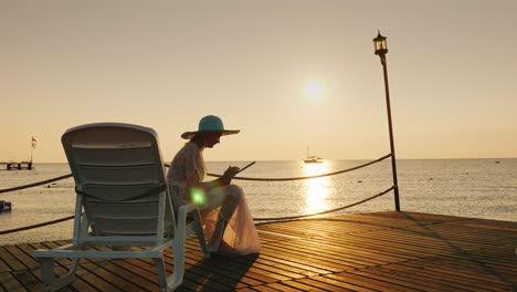 a woman in summer clothes sits on a lounger on the pier. uses a tablet. a beautiful dawn on the sea, in the distance is seen a fishing boat