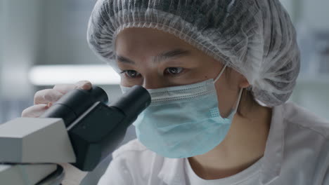 asian female scientist in mask observing sample through microscope