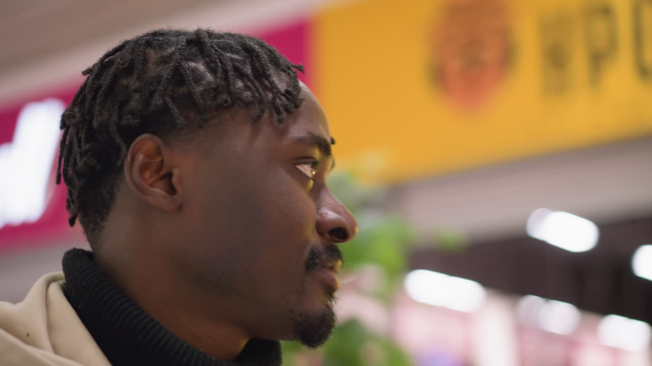 close up portrait of young black man with dreadlocks wearing hoodie and denim jacket looking around vibrant retail store interior with pink signage and bokeh lights capturing thoughtful urban shopper expression