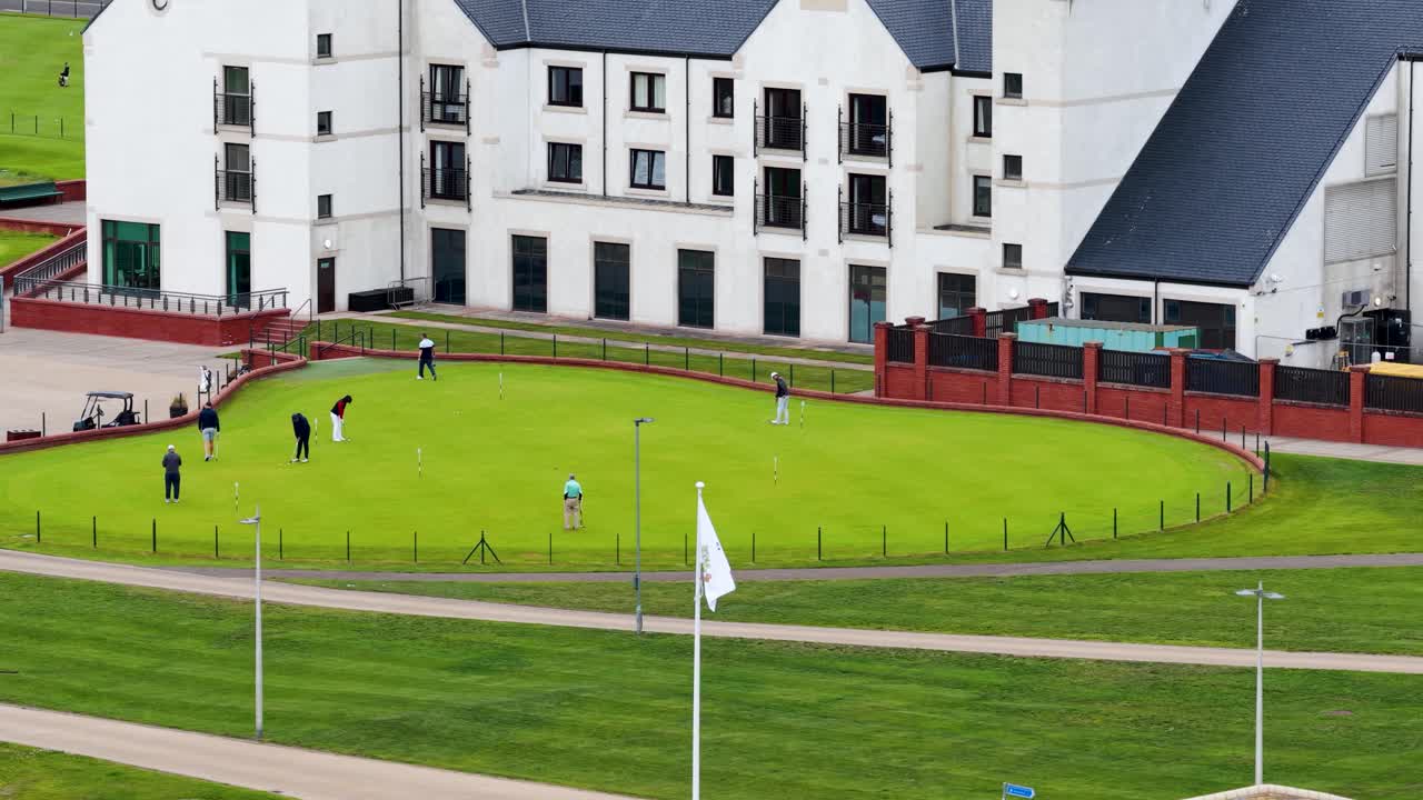 Golfers practice putting on green near modern clubhouse, captured by drone on bright summer day