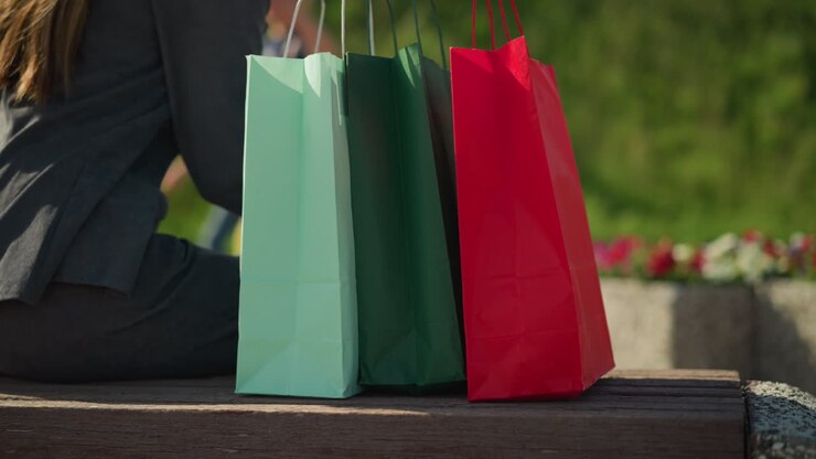 Close-up of an individual dropping colorful shopping bags on a bench outdoors, and sit down with a blurred partial view of two people walking in the background, flowers slightly visible