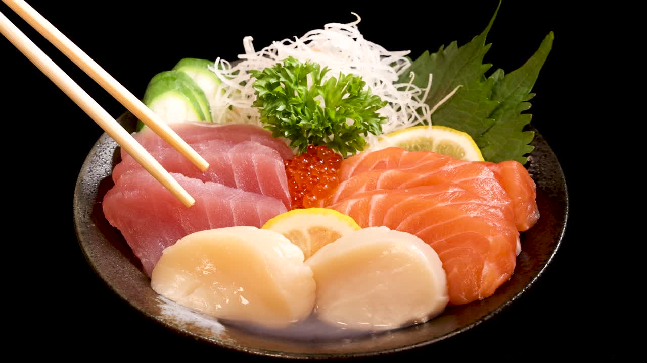 Chopsticks select scallop sashimi from a vibrant sushi set with salmon, tuna, roe, and garnishes on a black background under bright, even lighting
