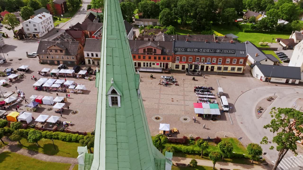 Drone rises around green church spire as market stalls fill Dobele square below