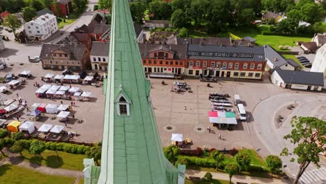 drone rises around green church spire as market stalls fill dobele square below