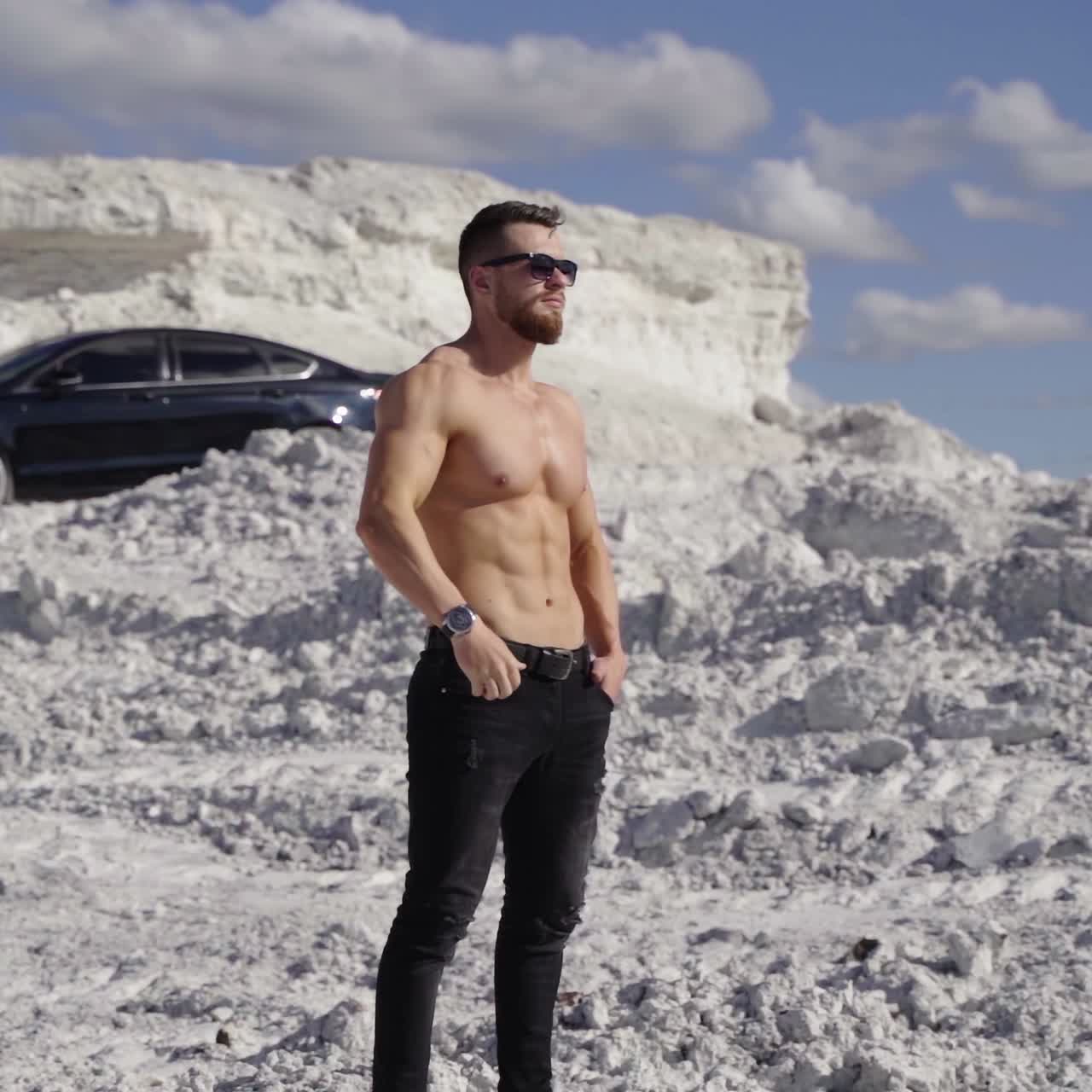 Shirtless athlete on white canyon. Portrait of handsome man with trained body in the mountains under the blue sky. Black car on the hill at sunny day.