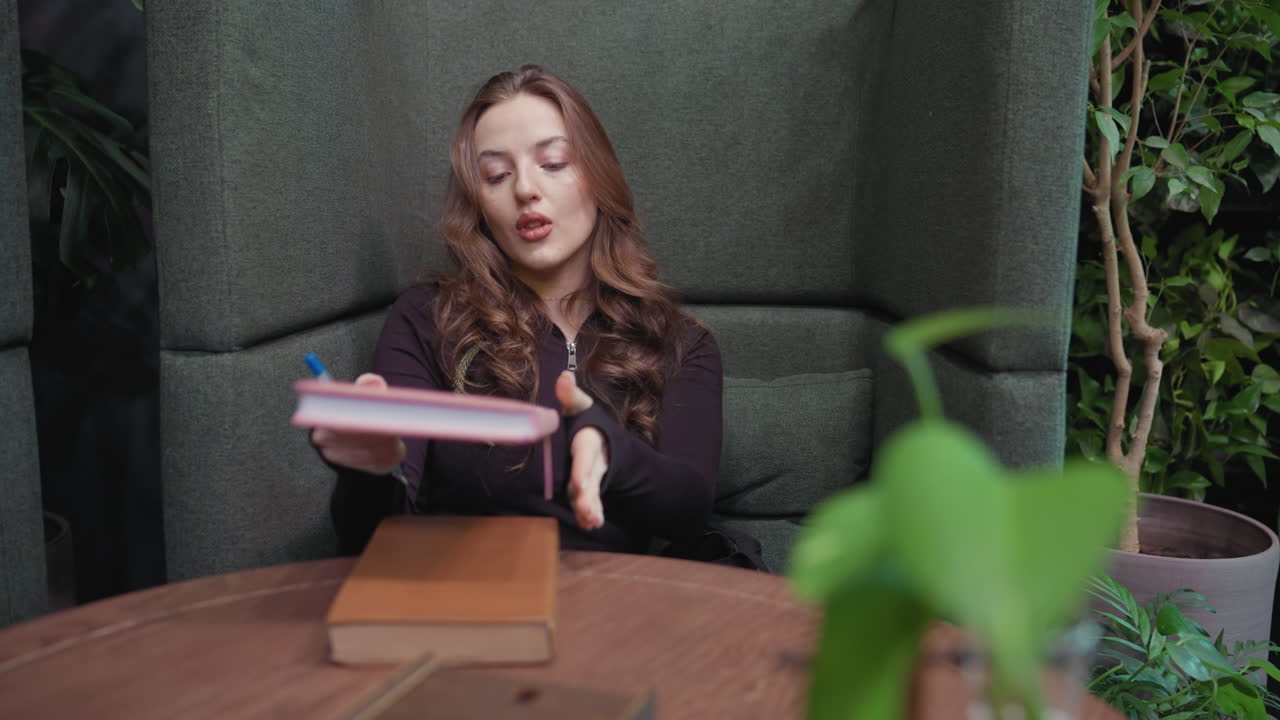 Blonde woman in black top sits quietly in green cushioned booth holding pink and brown books, with calm expression on face and indoor greenery in background adding natural serene atmosphere