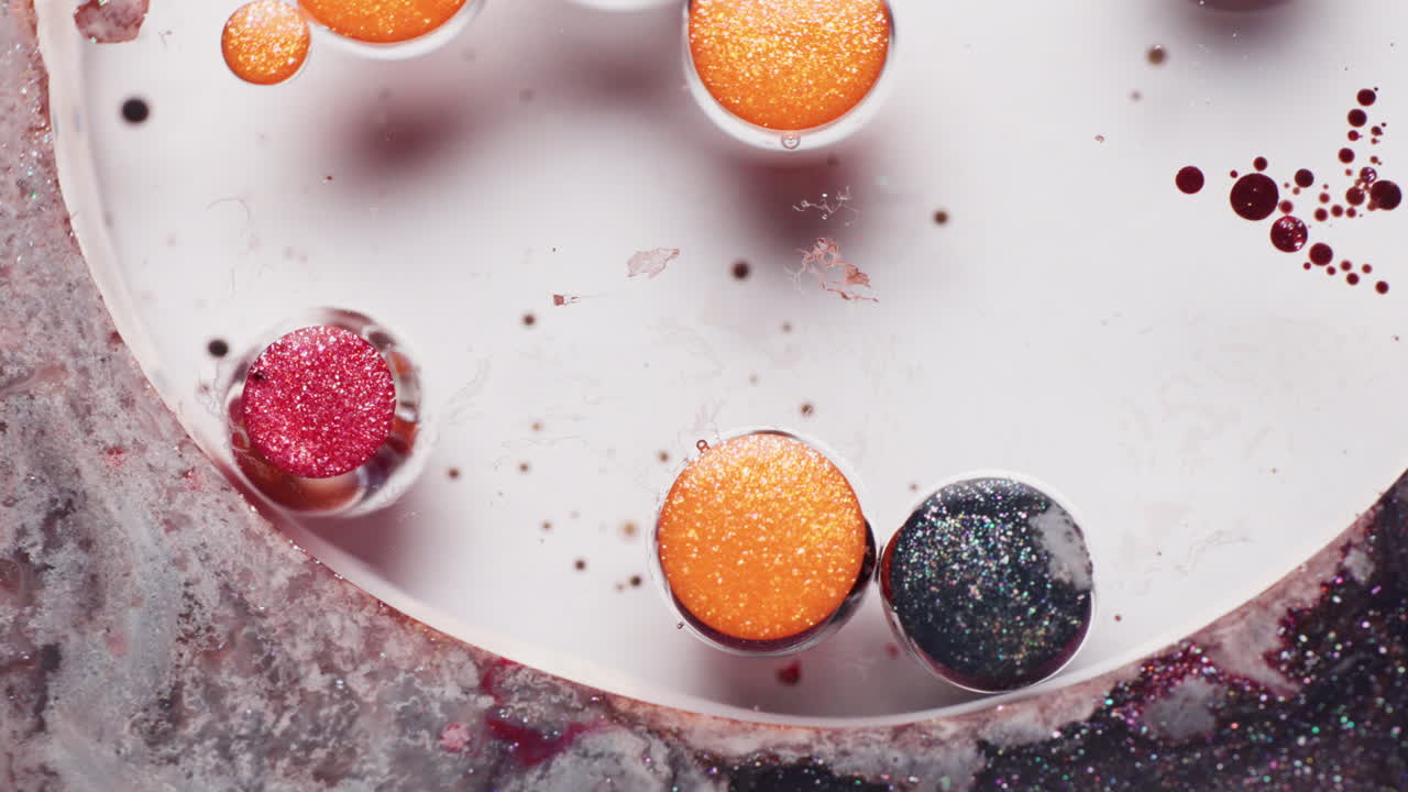 Macro zoom-in on red, orange, and black glitter ink droplets vibrating in a liquid solution with grey shimmering pigments around