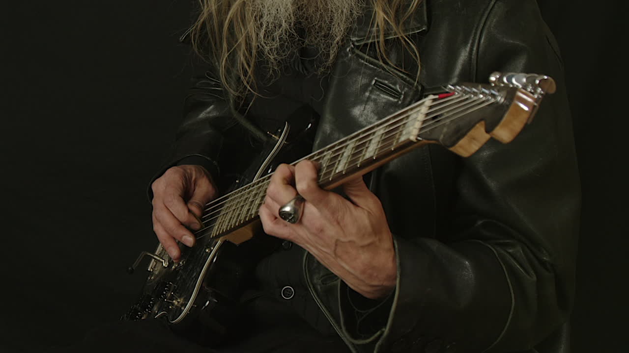Closeup: Bearded man in leather jacket plays electric guitar, dark room