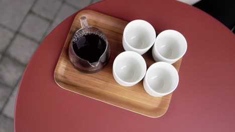 glass carafe and four white cups are arranged on wooden tray on red table