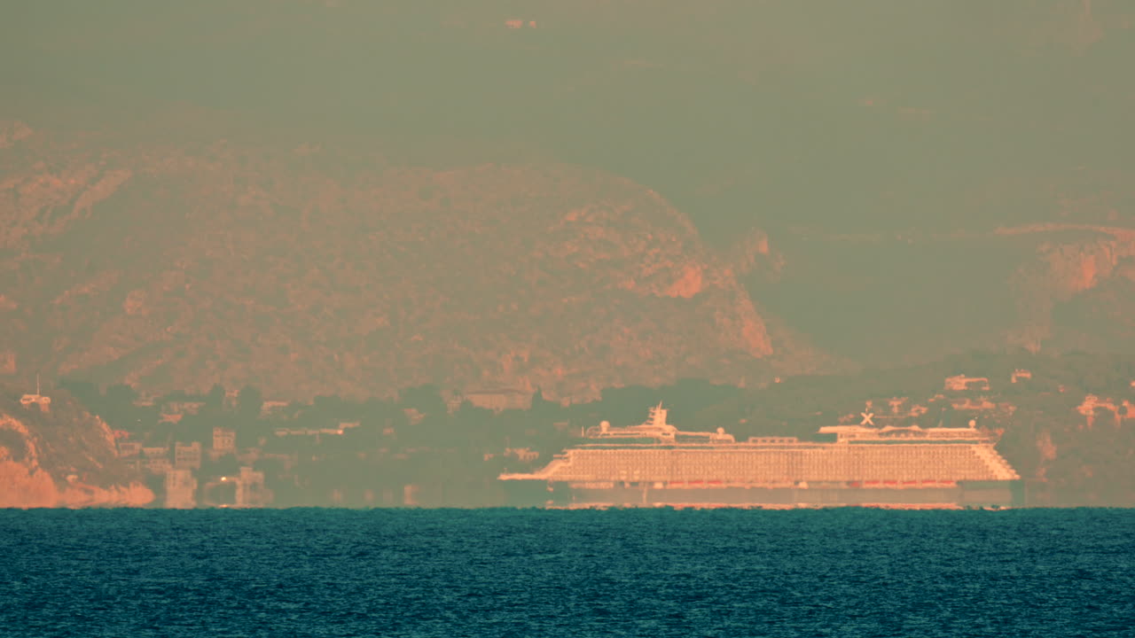 A distant view of a massive cruise ship sailing along the coastline, framed against hazy mountains and soft golden light