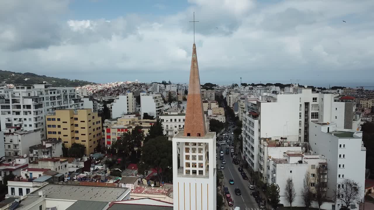 Tomada cinematográfica de drone de um campanário de igreja com vista para uma rua movimentada da cidade e edifícios brancos em Tânger, Marrocos, com o mar no horizonte.