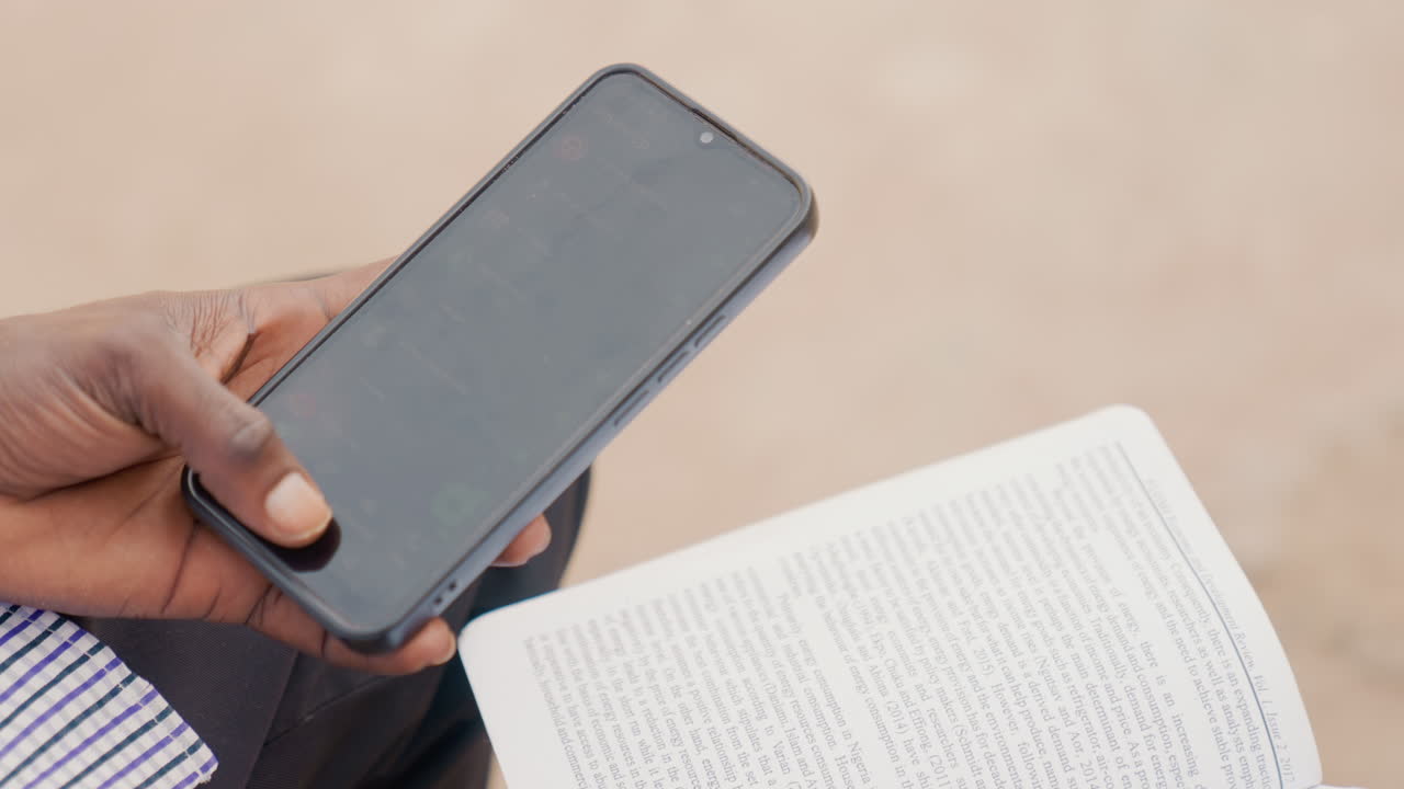 Uomo di colore che tiene telefono e quaderno, concentrato sugli appunti di studio all'aperto, primo piano delle mani che sfogliano le pagine, manica di camicia a righe, schermo dello smartphone fioco, testo scritto a mano visibile, atmosfera calma e concentrata.