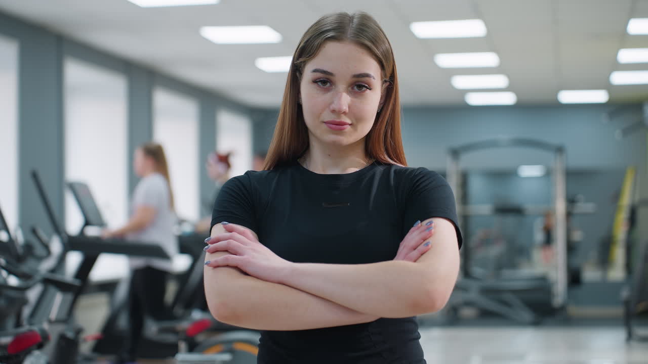 Confident elegant woman in black outfit posing with arms crossed inside modern gym while people work out in background using treadmills and equipment under bright indoor lighting