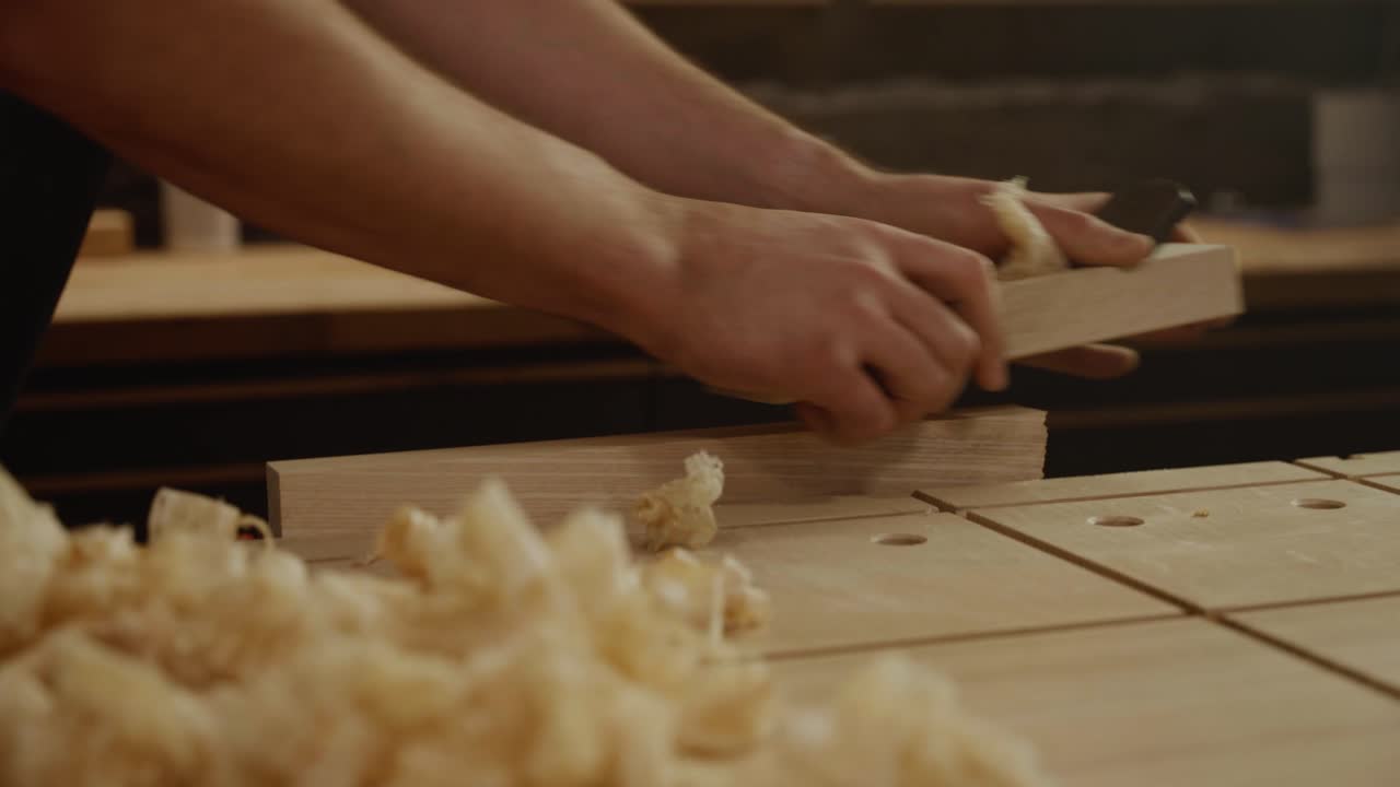 Close-up of a craftsman’s hands planing a wooden board with wood shavings scattered on the workbench, symbolizing craftsmanship, precision, and traditional woodworking