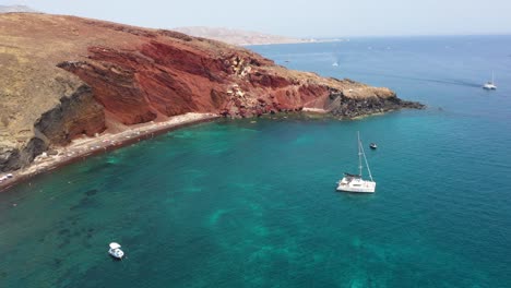 Giro-Aéreo-Sobre-Playa-Roja-Con-Agua-Turquesa,-Montañas,-Botes-Y-Arena-De-Color-Rojo-En-Santorini,-Grecia