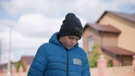 primer plano de un niño en chaqueta azul y gorra negra caminando al aire libre pensativo, el fondo borroso presenta una casa de ladrillo y un suave cielo de otoño