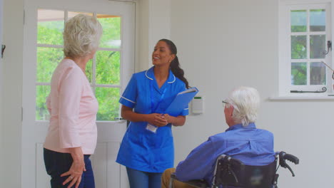 senior couple with man in wheelchair greeting female nurse or care worker making home visit at door