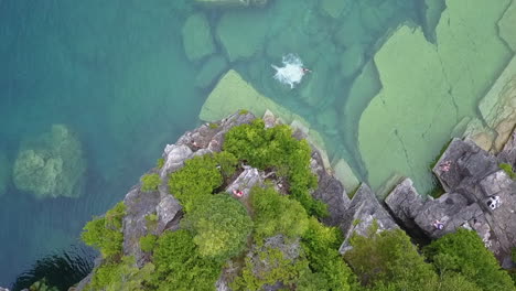 young man jumps off cliff into clear green lake water far below