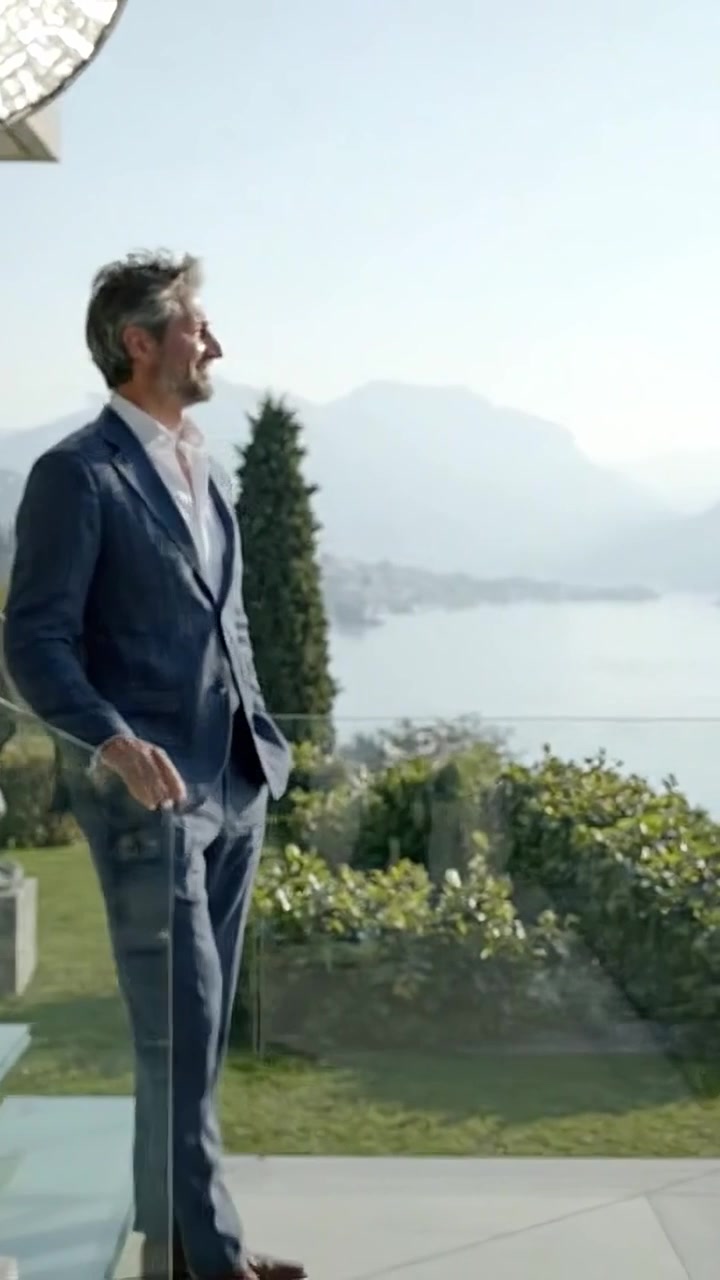 Un uomo in abito sta su un balcone di vetro che si affaccia su un lago sereno circondato da montagne, enfatizzando eleganza e tranquillità in un ambiente mozzafiato.