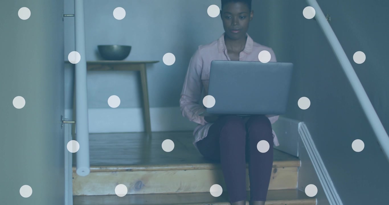 Animazione di macchie bianche su una donna afroamericana che usa un laptop sulle scale di casa.