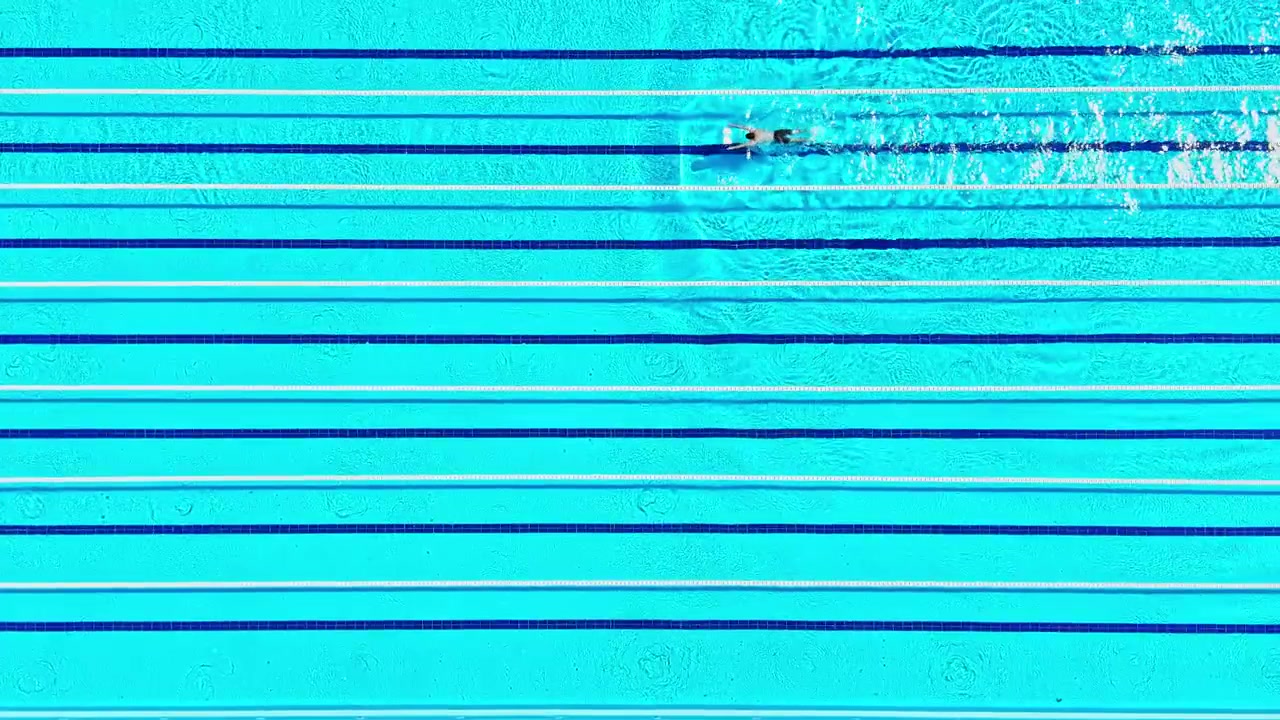 Seseorang berenang di kolam renang besar dengan jalur biru yang ditandai di air di bawah cahaya siang hari yang cerah.