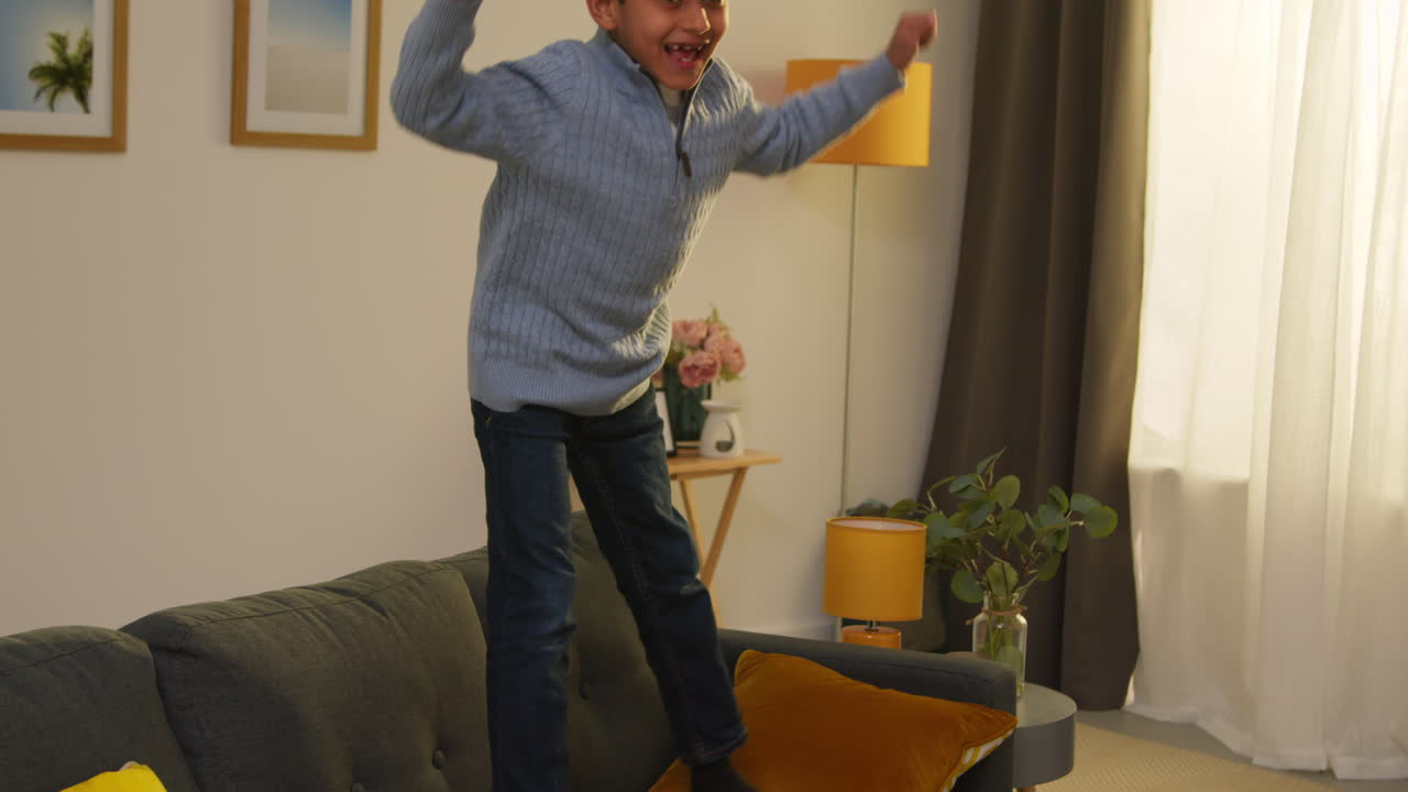 Disruptive Young Boy Behaving Badly At Home Jumping On Sofa Lounge