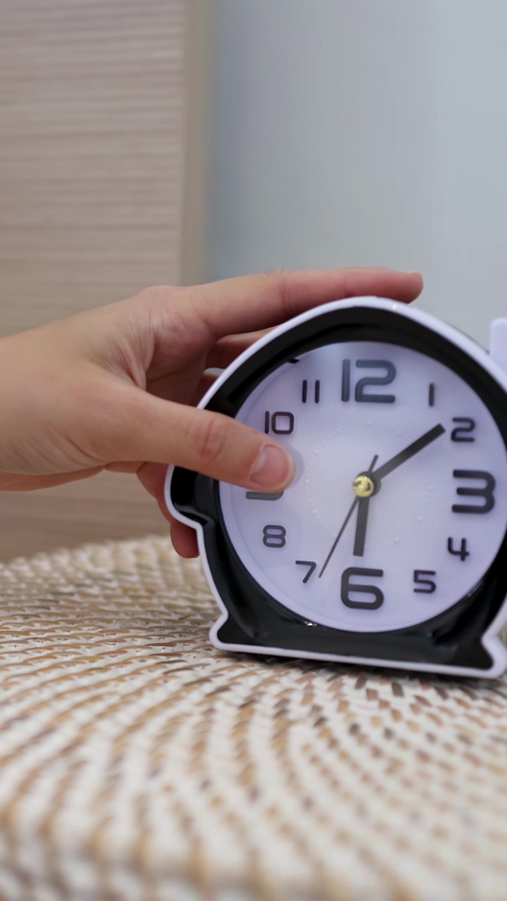 Una mano posa una sveglia analogica in bianco e nero su un comodino intrecciato