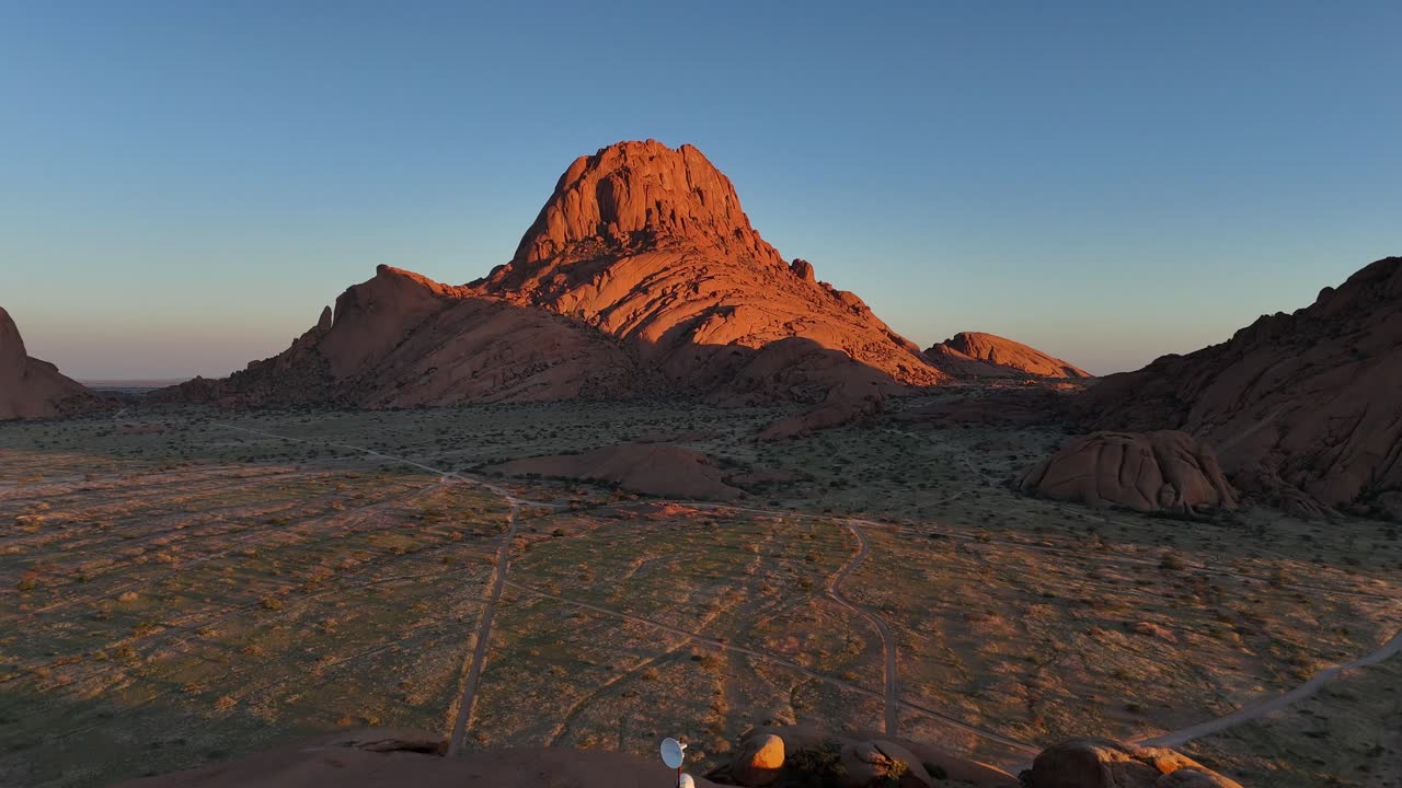 Aerial drone view of Spitzkoppe mountain in Namibia at golden hour, showing warm light over granite peaks and desert plains under a clear blue sky