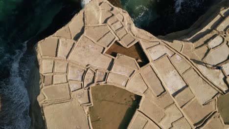 Aerial-high-angle-tracking-shot-of-a-man-walking-across-pools-of-salt-pans-in-Xwejni-during-a-beautiful-sunset