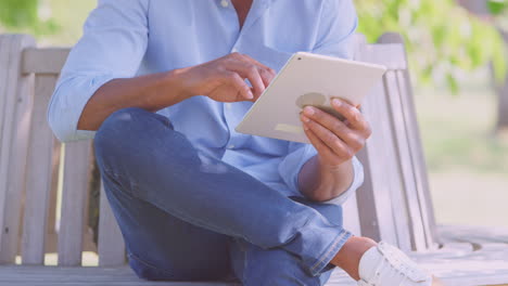 close up of man sitting on bench under tree in summer park using digital tablet