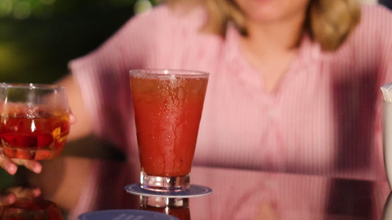 Donna in camicia rosa che prende un bicchiere da cocktail su un tavolo all'aperto illuminato dal sole, profondità di campo ridotta